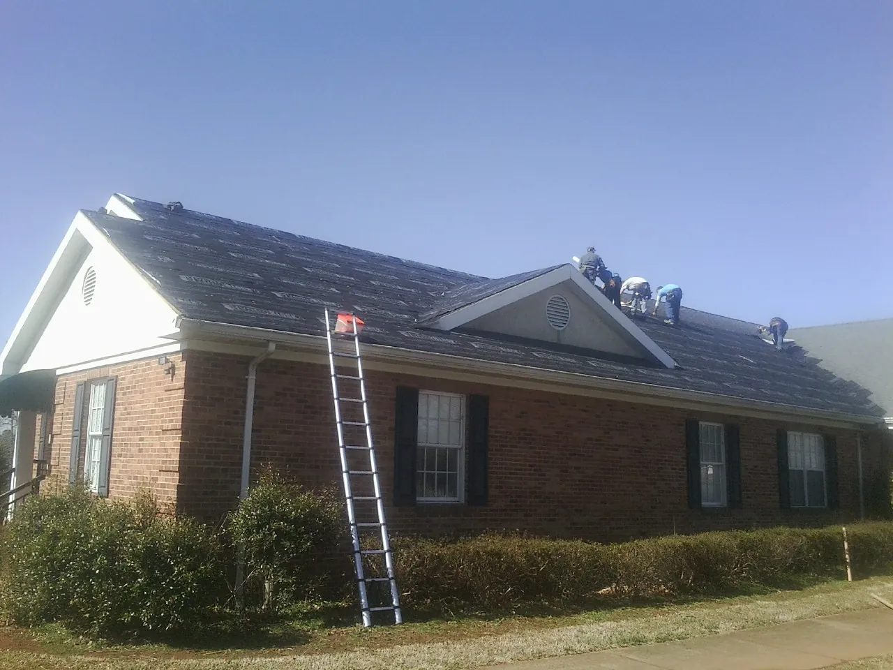 Crew tearing shingles off a brick ranch roof with a ladder set — Emergency Roof Repair work by our crew in Hallam