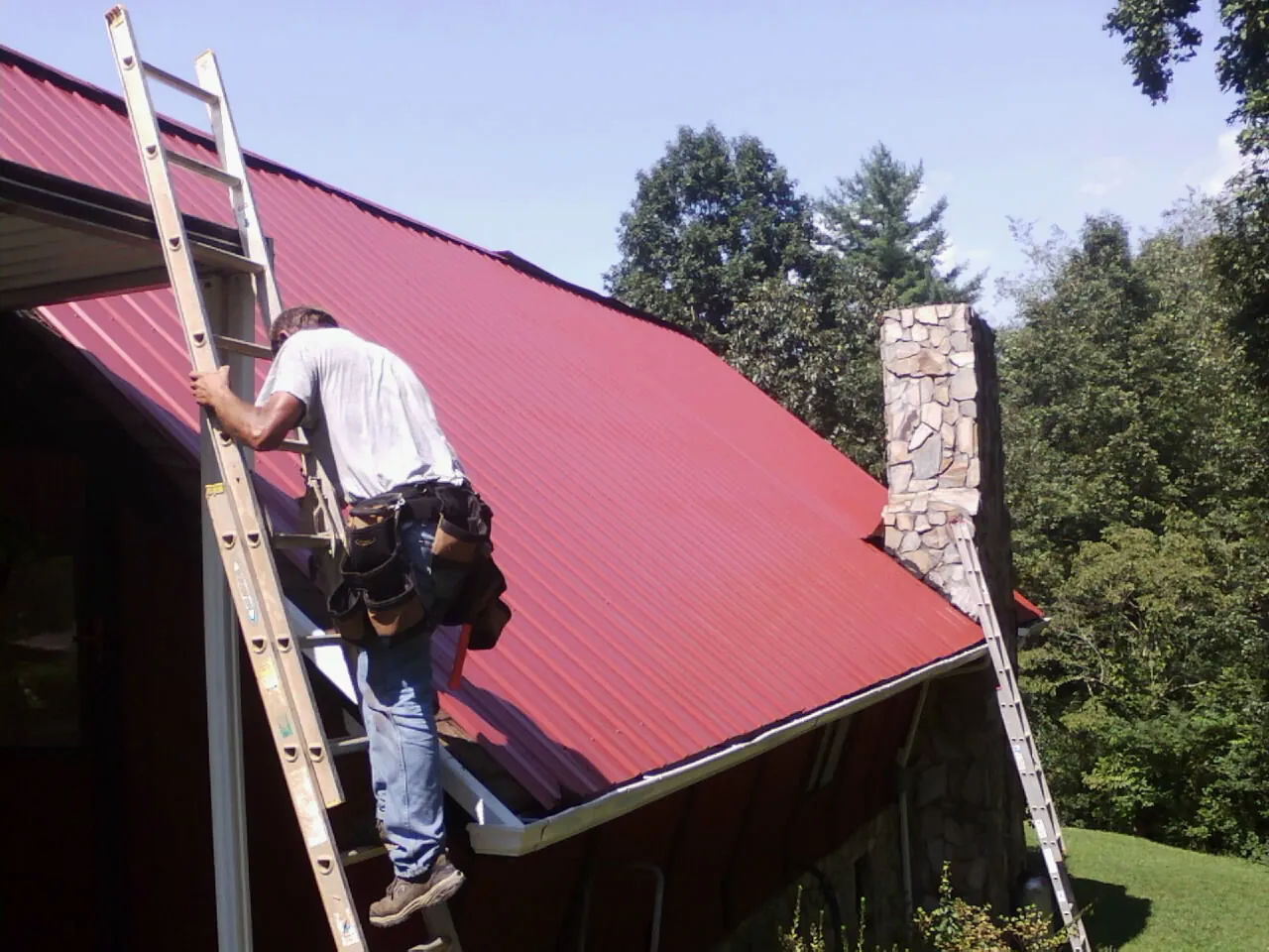 Roofer on a ladder beside a red metal roof and stone chimney — Soffit & Fascia Repair by Roofing Brothers in Hallam